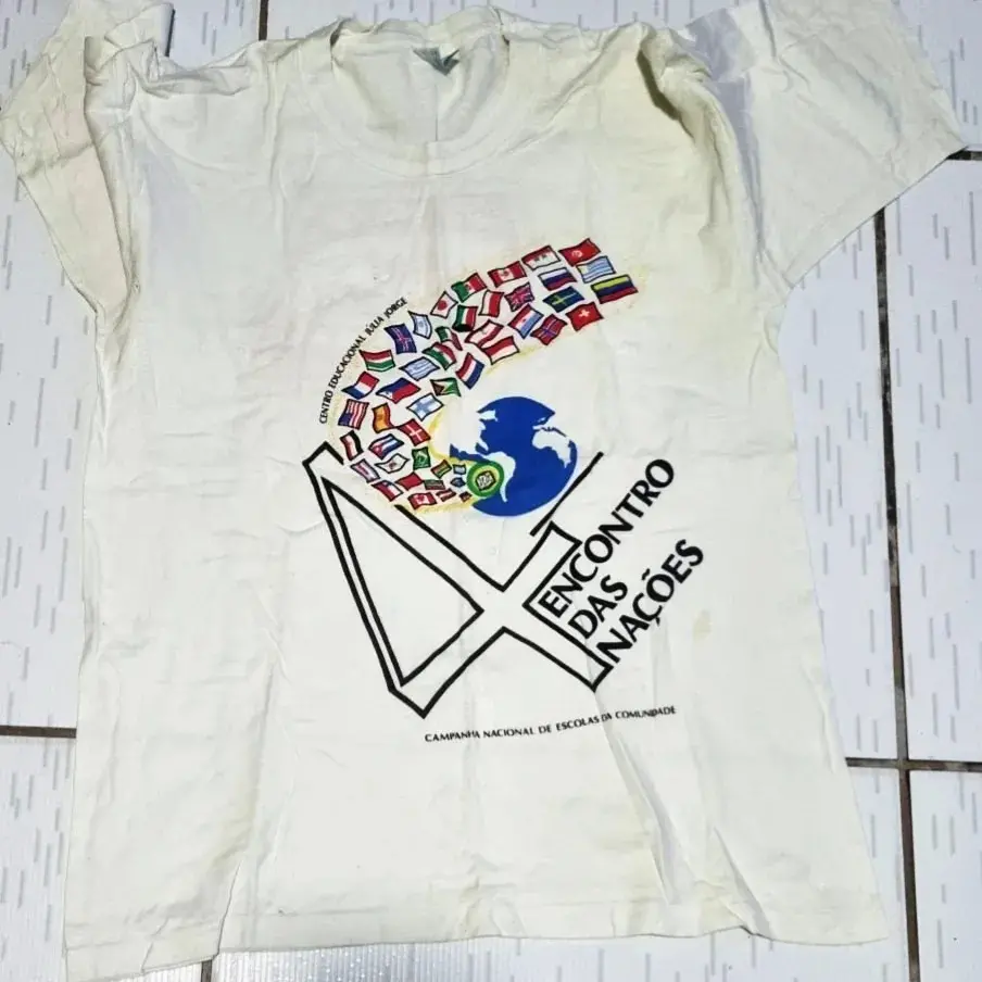 Camisa branca antiga estendida sobre um piso de ladrilhos claros. A camisa apresenta uma estampa no centro com as palavras ENCONTRO DAS NAÇÕES e um desenho de uma cruz de contorno preto, um globo azul e diversas bandeiras de países coloridas. A frase CAMPANHA NACIONAL DE ESCOLAS DA COMUNIDADE aparece abaixo do desenho.