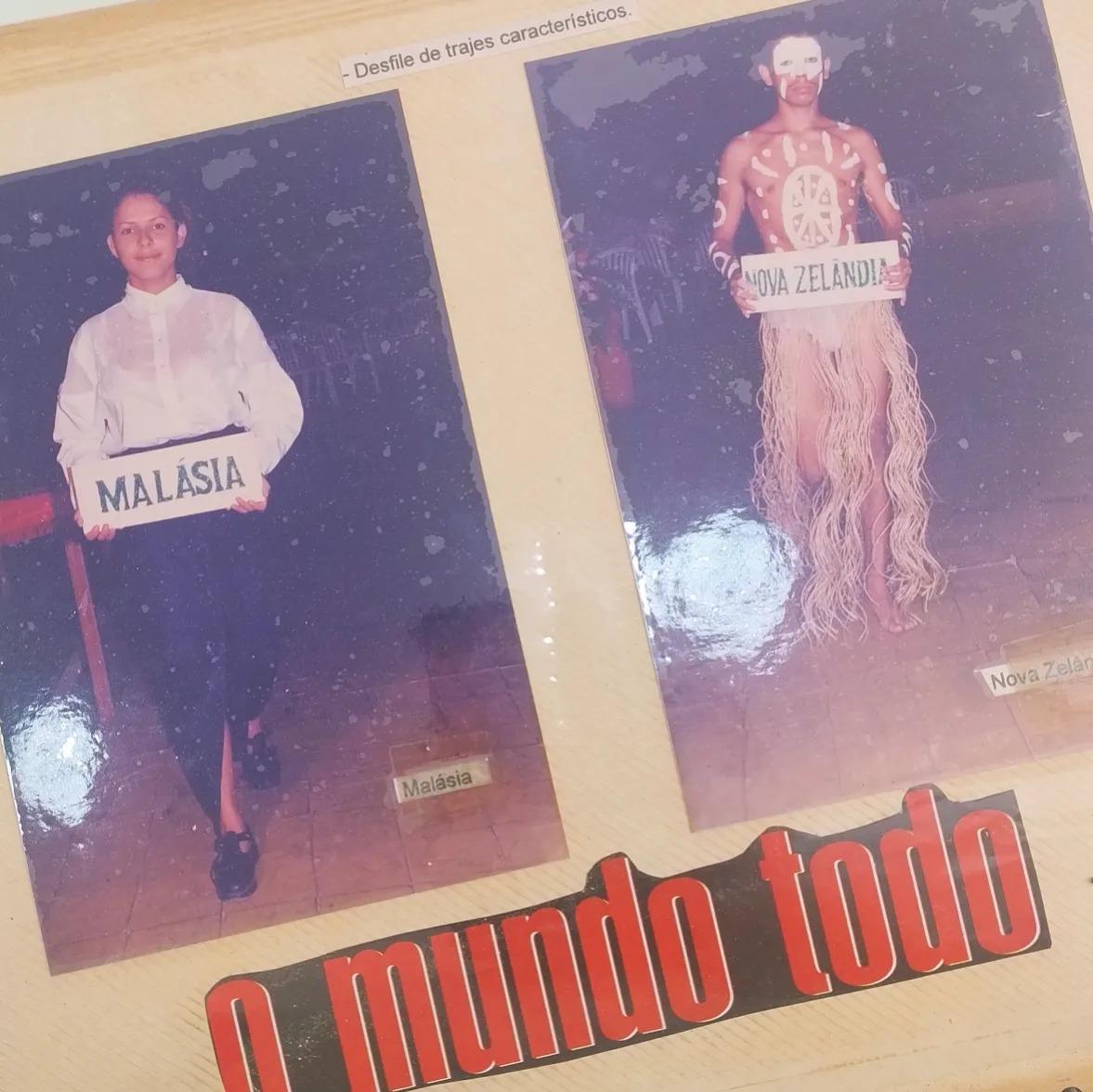Duas fotos antigas de um evento escolar, fixadas em um painel. A foto da esquerda mostra uma garota com camisa branca e saia escura segurando uma placa MALÁSIA. A foto da direita mostra um garoto com pintura corporal, saia de fibras e uma placa NOVA ZELÂNDIA.