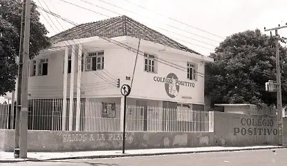 Foto em preto e branco de um prédio de dois andares com telhado de telhas, possivelmente um colégio, em uma esquina urbana. Um letreiro na fachada superior direita exibe COLÉGIO POSITIVO e seu logotipo. Há árvores e postes de rua visíveis.