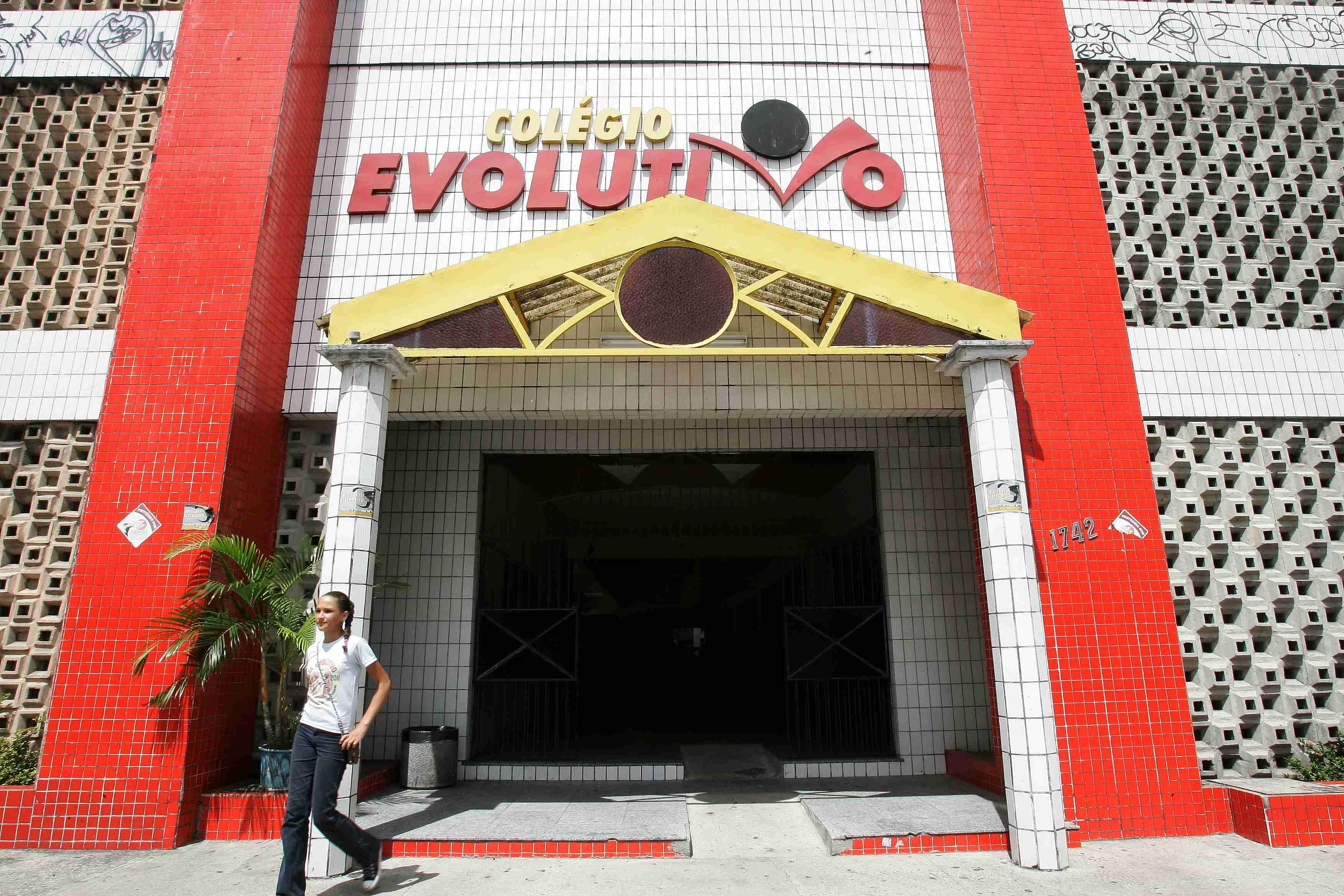 Foto da fachada do Colégio Evolutivo, em cores vibrantes. A entrada tem uma pequena marquise amarela sustentada por colunas brancas. O nome COLÉGIO EVOLUTIVO está em vermelho na parte superior, em destaque. As laterais têm paredes de tijolos vazados e pilares vermelhos. Uma jovem sorridente está no canto inferior esquerdo.