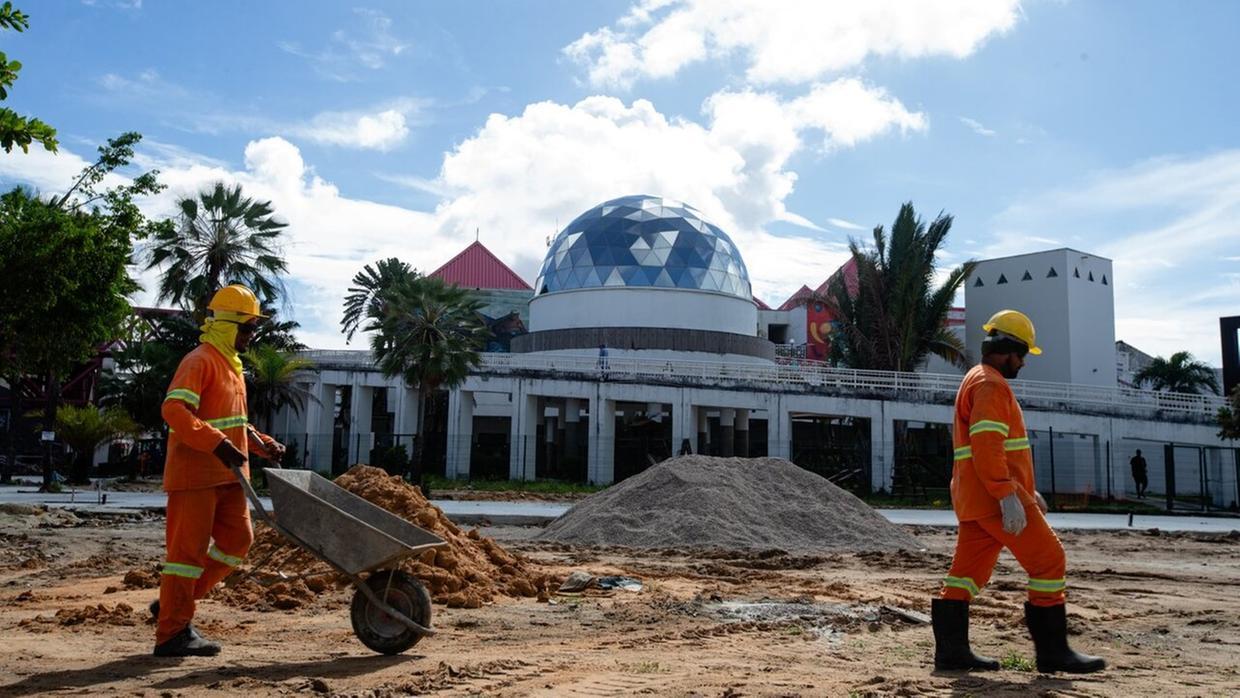 Na imagem, cena de construção ou manutenção em frente ao Centro Cultural Dragão do Mar em Fortaleza, Ceará. Em primeiro plano, dois operários vestindo uniformes de segurança laranja fluorescente e capacetes amarelos estão trabalhando em um terreno de terra batida. Um operário empurra um carrinho de mão e o outro caminha ao lado. Há um monte de areia e terra solta entre eles. Ao fundo, o complexo do Dragão do Mar é visível, destacando-se a cúpula geodésica azul e branca do planetário. A arquitetura de cor clara tem detalhes em telhado vermelho. Palmeiras e vegetação tropical emolduram a cena sob um céu azul claro com nuvens esparsas e brilhantes.