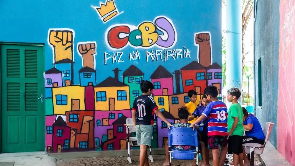 Na imagem, um grupo de crianças reunidas em frente a um mural vibrante de graffiti que cobre uma parede azul brilhante. O graffiti apresenta a sigla 