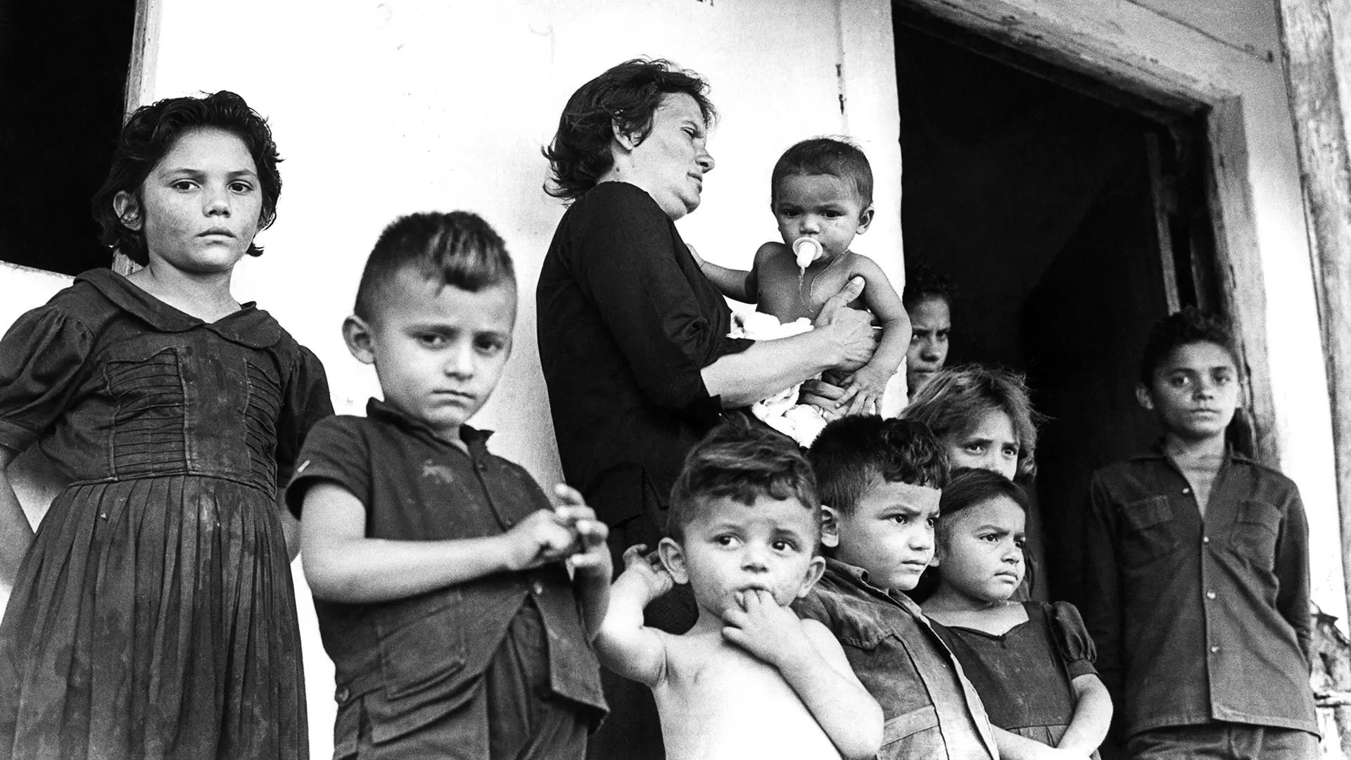 Cena do documentário 'Cabra Marcado Para Morrer', de Eduardo Coutinho. Foto em preto e branco de um grupo de crianças e uma mulher segurando um bebê, próximos de uma porta e com expressões sérias.