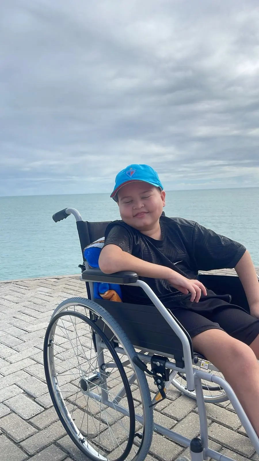 Criança feliz em cadeira de rodas na orla do mar com céu nublado ao fundo, aproveitando o dia ao ar livre perto da água.