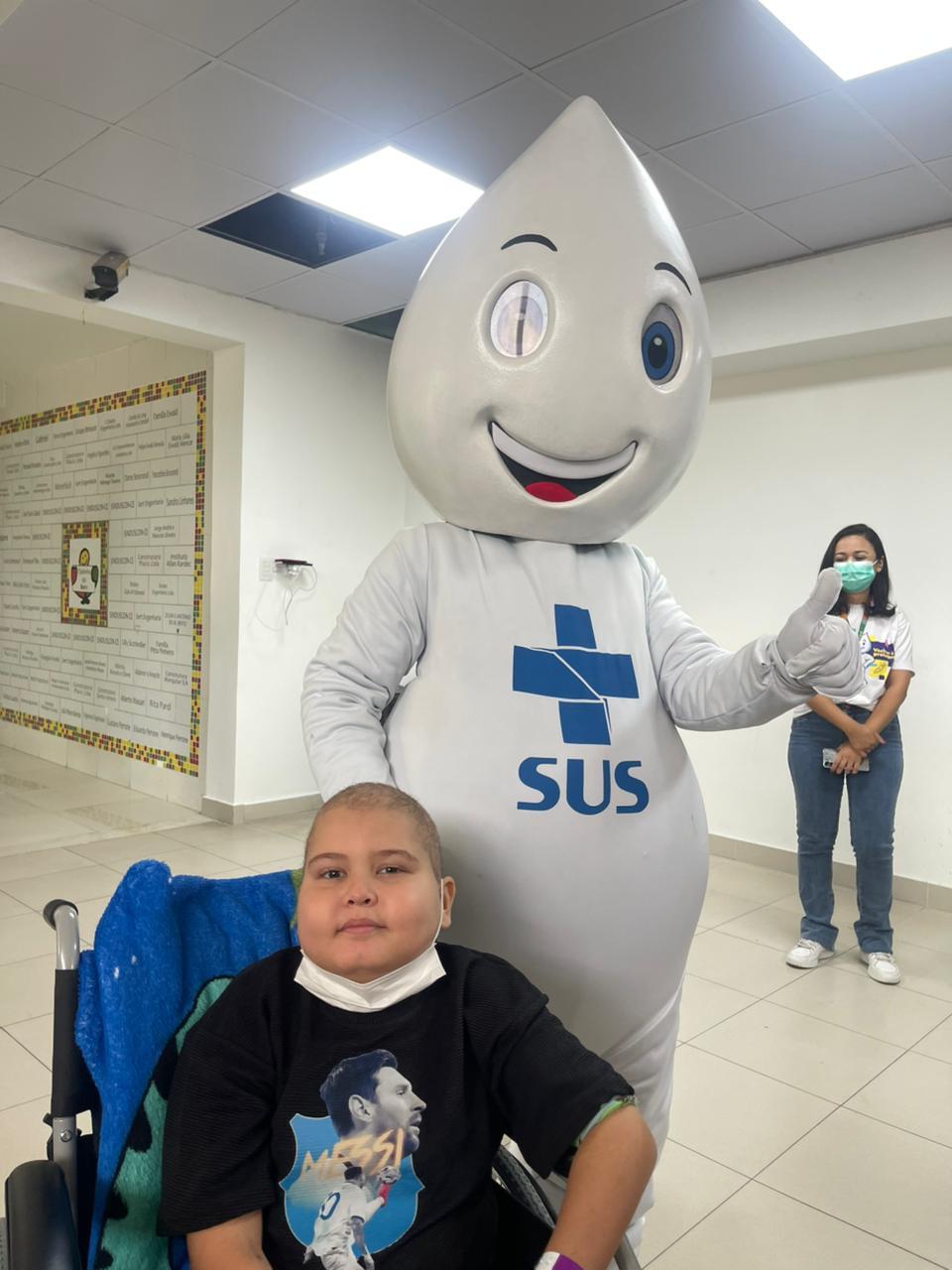 Criança com câncer deitado em cadeira de rodas, ao lado de mascote do SUS, representante do Sistema Único de Saúde, em ambiente hospitalar, enquanto uma mulher de máscara ao fundo observa.