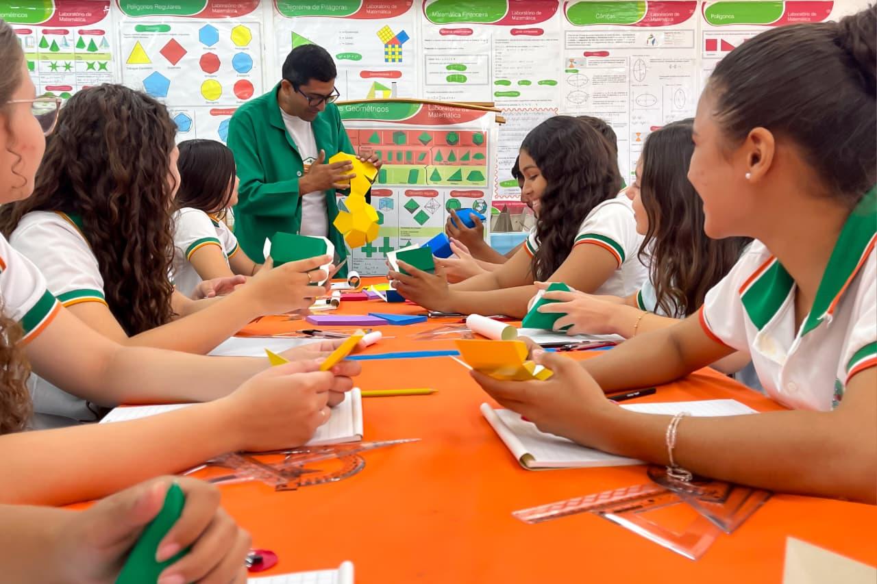 Em uma sala de aula de Matemática, um grupo de estudantes de uniforme está em torno de uma mesa. Eles manipulam modelos geométricos em verde e amarelo. Um professor com um jaleco verde está de pé no centro, interagindo com os alunos. A parede de fundo tem cartazes coloridos com temas geométricos.