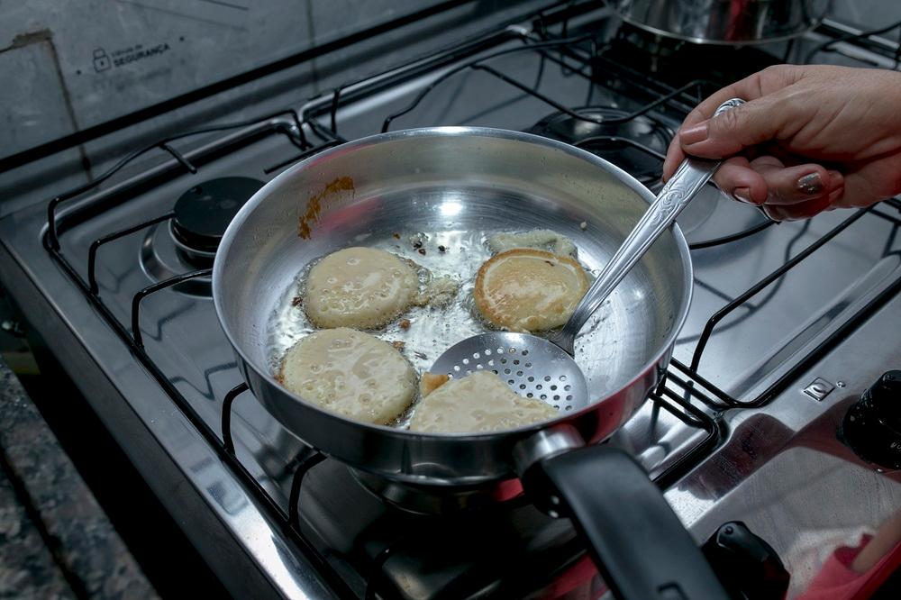 Na imagem, uma foto de close-up mostra uma panela de metal sobre um fogão a gás. Dentro da panela, há óleo quente onde quatro unidades de bruaca, ou um tipo de bolinho/panqueca frito, estão sendo preparadas. A bruaca tem uma forma redonda e irregular, com uma cor dourada clara.