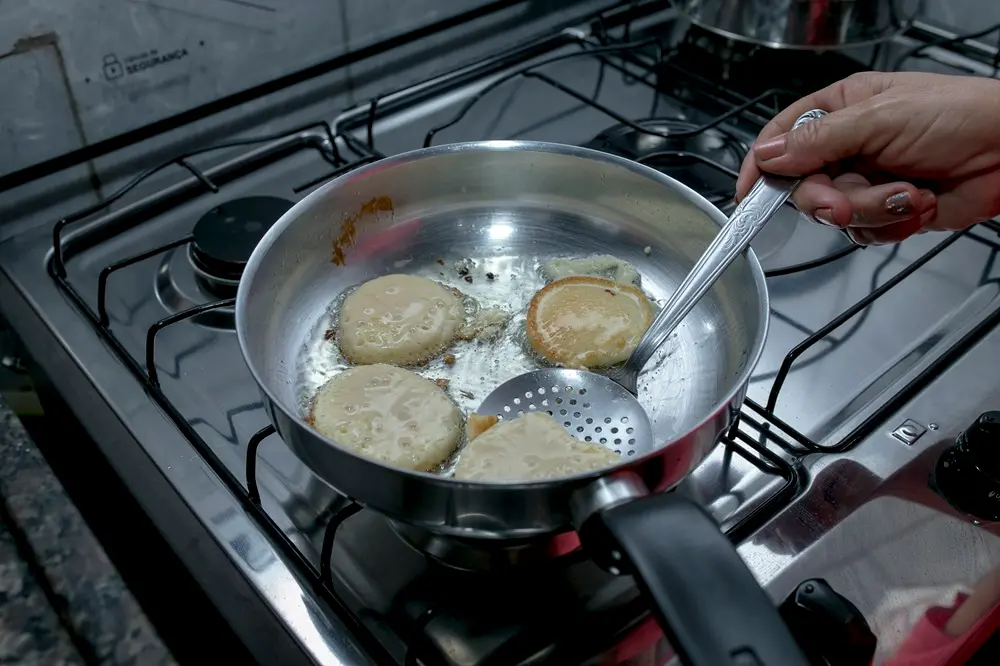 Na imagem, uma foto de close-up mostra uma panela de metal sobre um fogão a gás. Dentro da panela, há óleo quente onde quatro unidades de bruaca, ou um tipo de bolinho/panqueca frito, estão sendo preparadas. A bruaca tem uma forma redonda e irregular, com uma cor dourada clara.