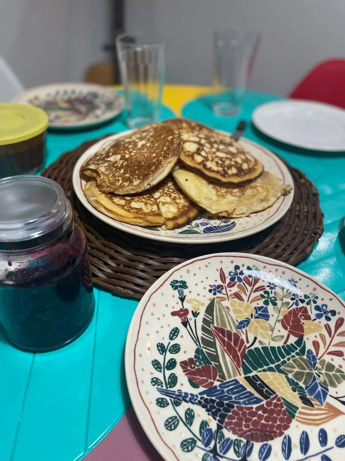 Na imagem, uma foto de cima e em close-up mostra uma pilha de bruacas (ou panquecas) em um prato decorado, que está apoiado sobre um jogo americano redondo e de textura trançada (possivelmente de vime ou palha). As bruacas são douradas, com bordas irregulares e manchas escuras da fritura ou chapa.