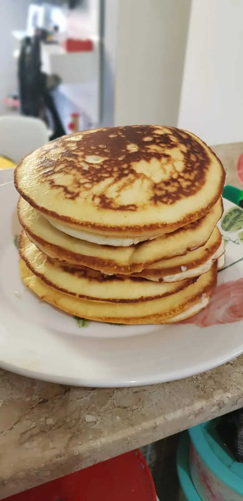 Na imagem, uma foto de close-up, com ângulo ligeiramente elevado, mostra uma pilha alta de quatro ou cinco bruacas (ou panquecas) em um prato branco com uma discreta decoração floral. As bruacas são redondas, grossas e fofas, com uma cor dourada nas bordas e nas laterais. O topo da pilha está bem bronzeado com manchas marrons mais escuras, indicando que foram bem cozidas.