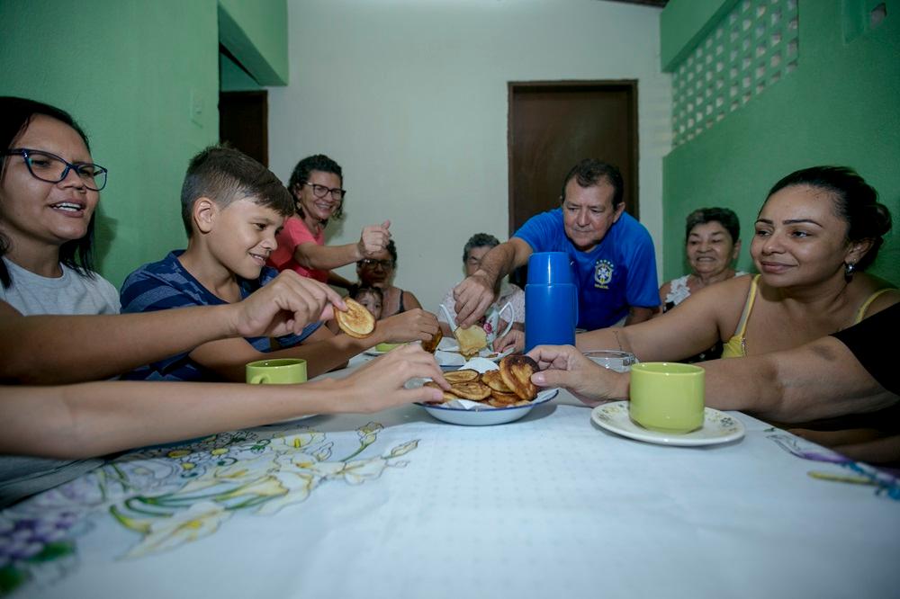 Na imagem, uma foto em plano médio captura uma família reunida em torno de uma mesa, desfrutando de uma refeição. Várias pessoas de diferentes idades, incluindo crianças, adultos e idosos, estão sentadas ao redor da mesa coberta por uma toalha branca estampada com flores amarelas. No centro da mesa, um prato com pequenas porções de bruaca (ou bolinhos/panquecas) é compartilhado. As mãos das pessoas estão estendidas para pegar os pedaços. Um homem de camisa azul, no centro, está distribuindo a comida.