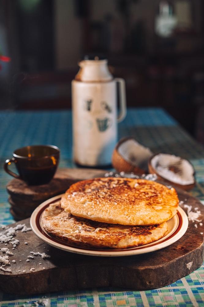 Na imagem, duas bruacas (panquecas grossas e douradas) empilhadas em um prato com bordas marrons, colocadas sobre uma tábua de madeira rústica. Em primeiro plano, há coco ralado espalhado. No fundo, fora de foco, há uma xícara de café escura sobre outro pedaço de madeira, um coquinho partido e uma garrafa térmica antiga. A mesa é coberta por uma toalha xadrez em tons de azul e verde.
