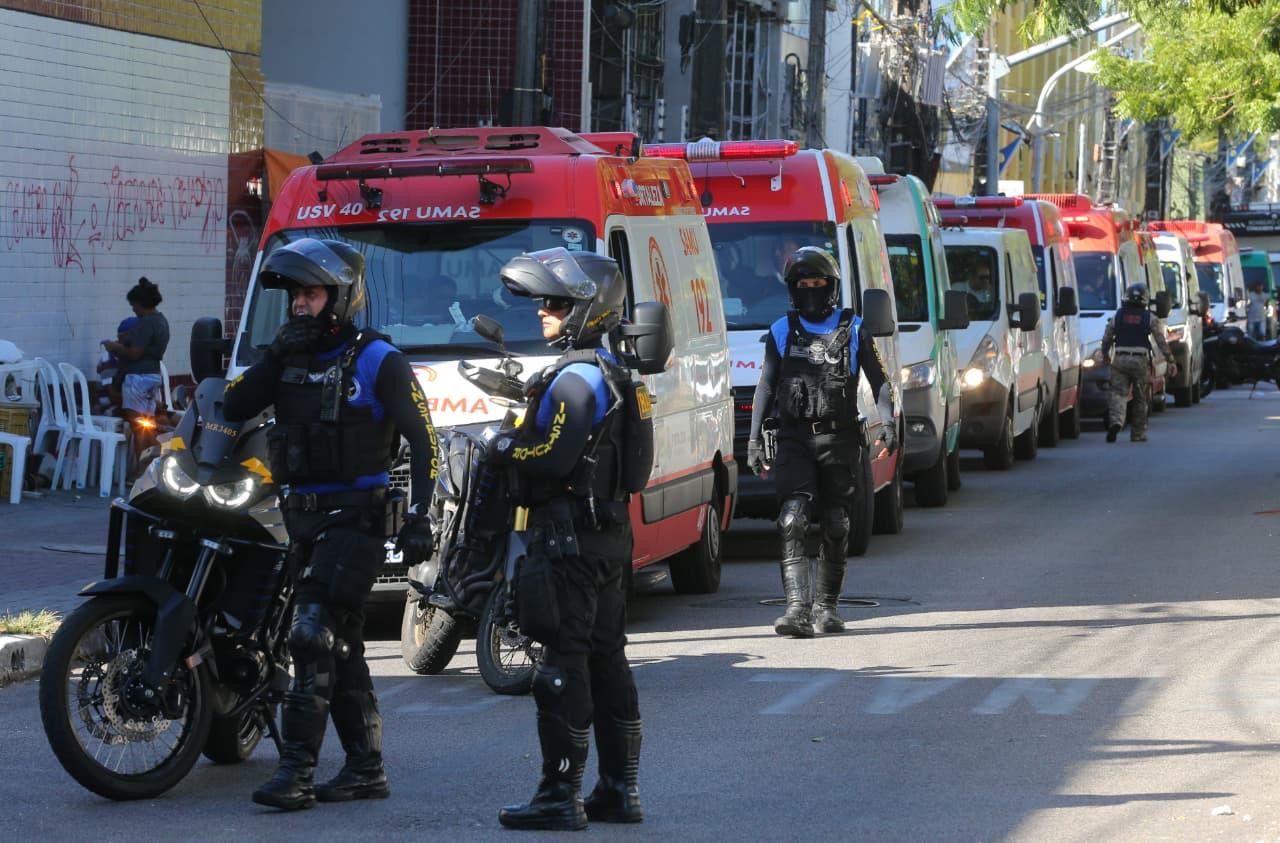 Agentes de segurança motorizados, usando capacetes e coletes táticos, circulam e vigiam uma rua onde várias ambulâncias do SAMU estão alinhadas. Ao fundo, moradores aparecem próximos a cadeiras brancas encostadas em uma parede grafitada.