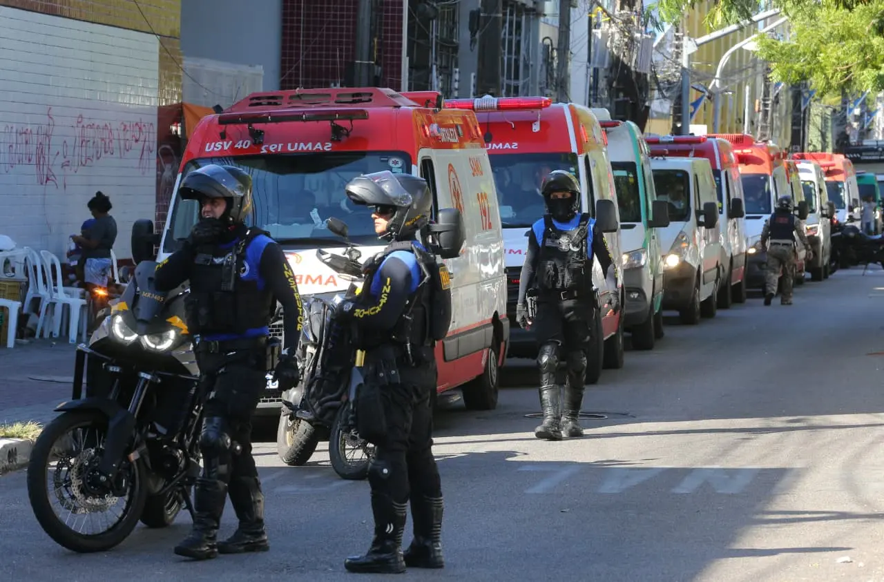 Agentes de segurança motorizados, usando capacetes e coletes táticos, circulam e vigiam uma rua onde várias ambulâncias do SAMU estão alinhadas. Ao fundo, moradores aparecem próximos a cadeiras brancas encostadas em uma parede grafitada.