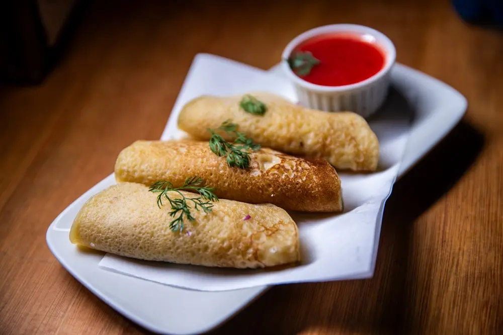 Na imagem, três bruacas douradas e enroladas, servidas sobre um guardanapo branco em um prato retangular. As bruacas estão guarnecidas com um raminho de endro ou coentro no topo e são acompanhadas por um pequeno recipiente branco com molho vermelho (geleia ou calda) ao fundo. O prato está sobre uma superfície de madeira escura.