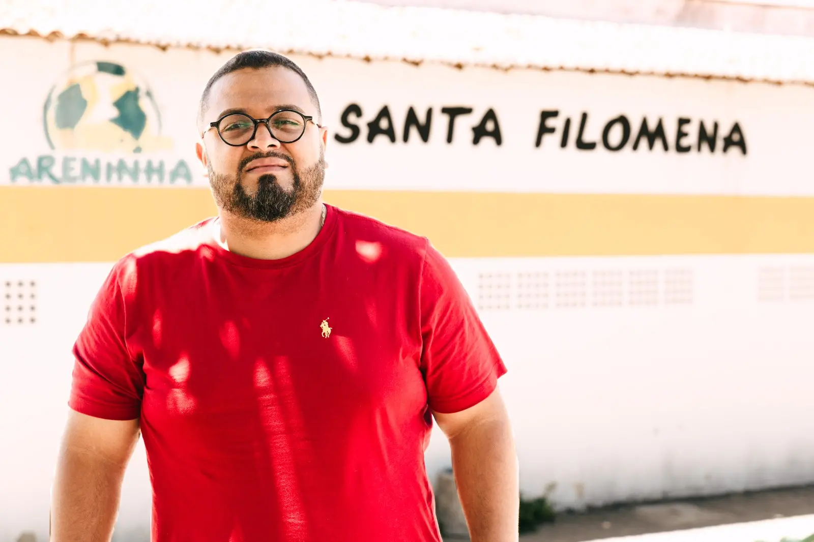 Retrato em plano médio de um homem de barba e óculos, vestindo uma camiseta vermelha, posando sob a luz do sol. O fundo é uma parede branca com uma faixa amarela onde se lê 'Areninha Santa Filomena' e um logo de bola/campo.