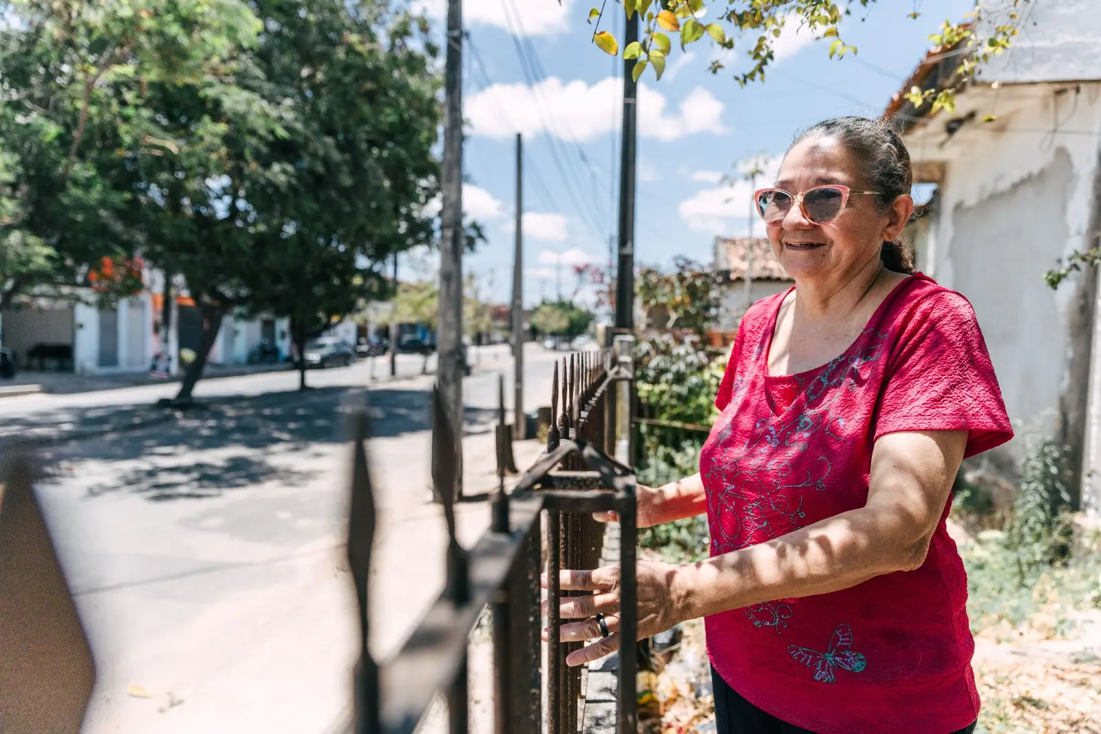 Retrato de uma mulher sorrindo, de óculos de sol rosa e camisa vermelha/rosa, apoiada em uma cerca de ferro. Ao fundo, uma rua ensolarada de bairro com árvores e postes de eletricidade..