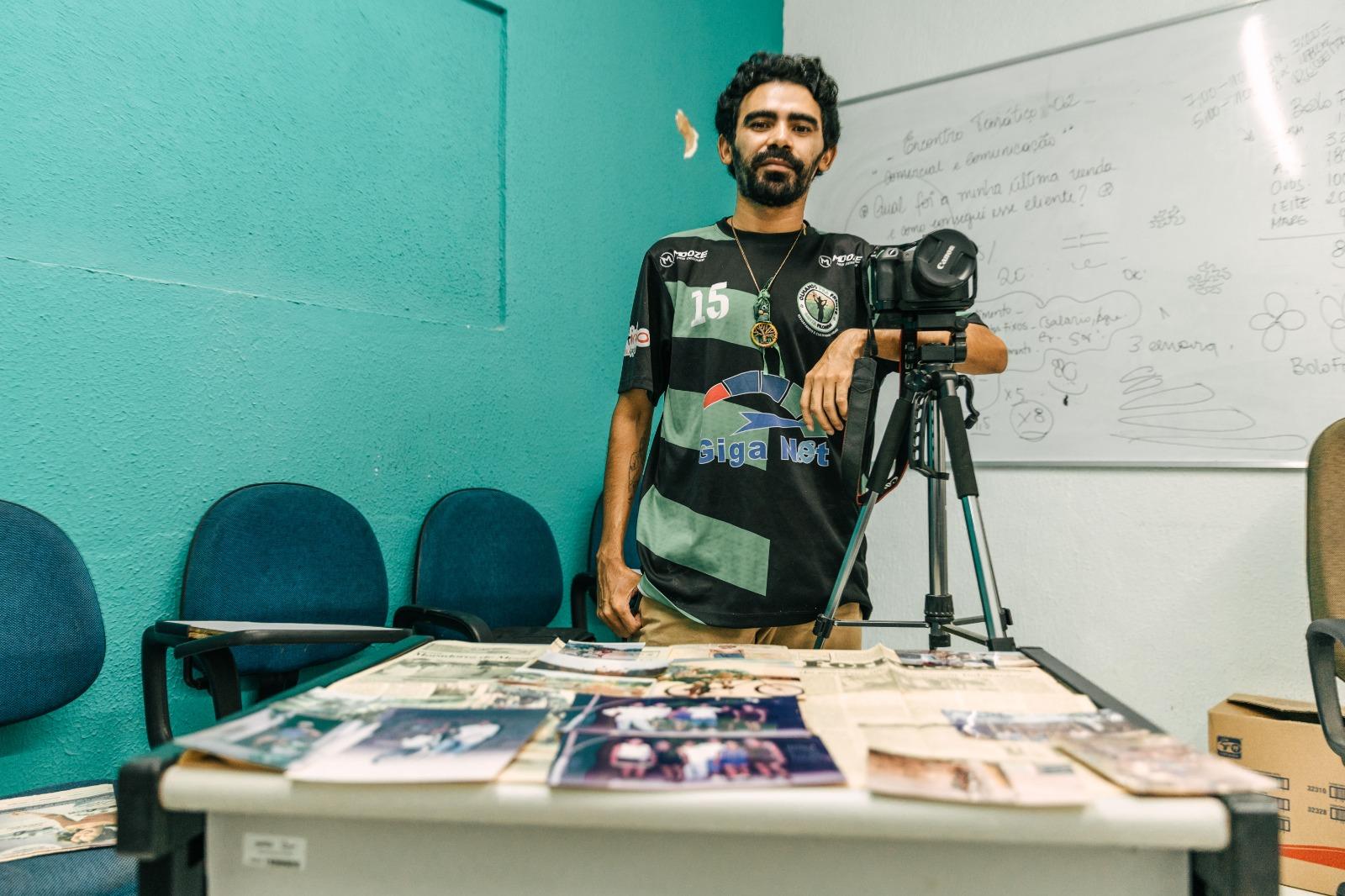 Retrato de um jovem de barba e de camiseta esportiva, apoiado em uma câmera em tripé, dentro de uma sala com paredes azul-turquesa e um quadro branco com anotações. Várias fotos impressas e jornais estão espalhadas em uma mesa à sua frente.