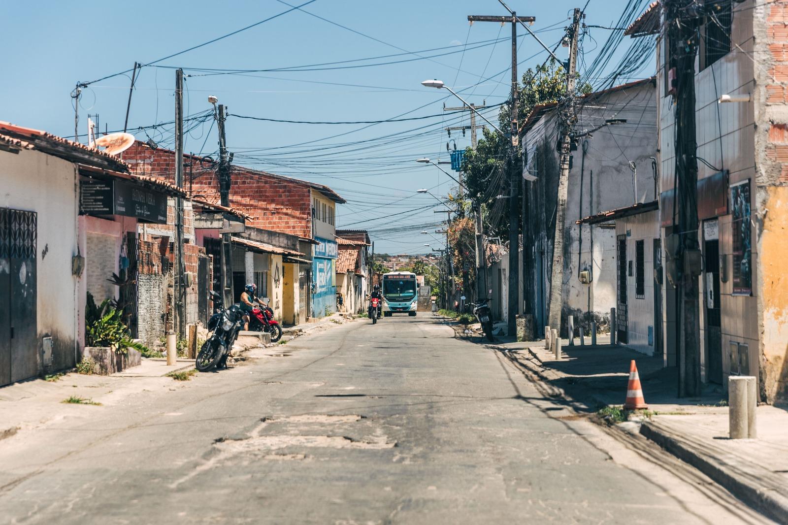 Fotografia de uma rua de bairro popular, estreita e ensolarada, ladeada por casas simples de tijolo e reboco. Muitos postes e uma densa rede de cabos cruzam o céu. Um ônibus branco e azul se aproxima ao longe, no centro da rua.