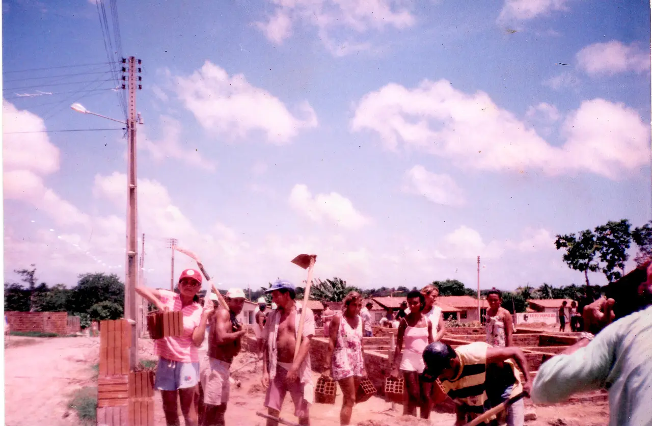 Fotografia de um mutirão de construção de moradias populares, com homens e mulheres segurando materiais de construção diversos. Postes e redes de cabos cruzam o céu.