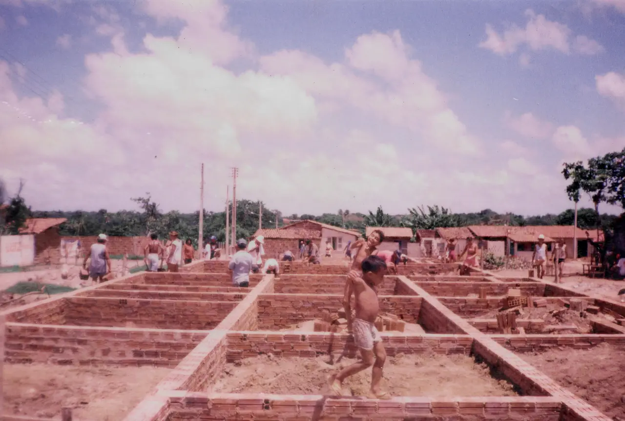 Foto antiga, com tons sépia/rosa, mostrando um canteiro de obras de moradias populares. Alicerces de tijolo formam o esqueleto de várias casas. No centro da foto, é possível ver dois meninos brincando na estrutura ainda em construção. Ao fundo, casas de tijolo e vegetação.