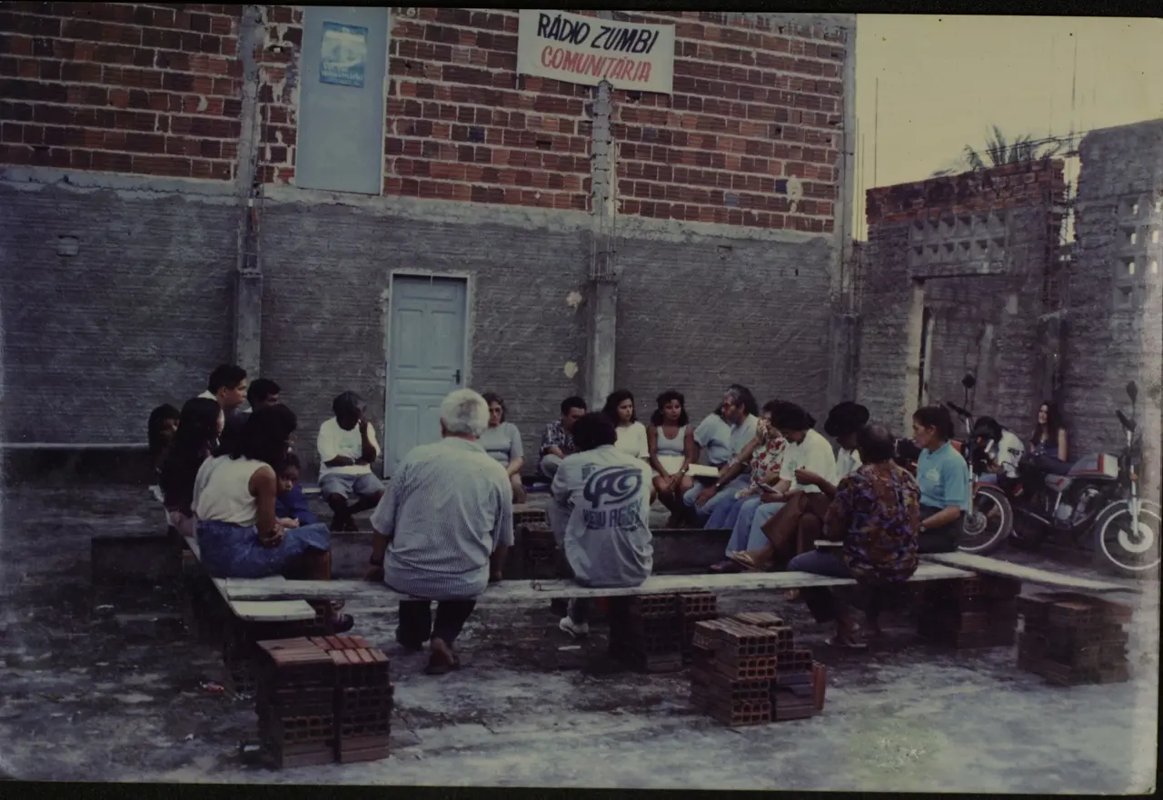 Foto antiga de um grupo de mais de 20 pessoas reunidas em bancos improvisados de madeira apoiados em tijolos. Eles estão em frente a um prédio rústico de tijolos e cimento. Uma faixa na parede diz 'Rádio Zumbi Comunitária'.
