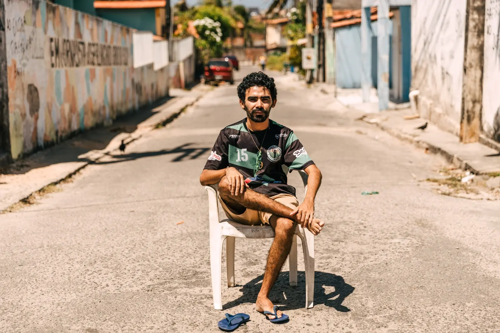 Retrato do cineasta Leo Silva, que usa barba e camiseta esportiva, sentado em uma cadeira de plástico branca no meio de uma rua de bairro. O muro à esquerda é grafitado. O sol forte projeta sombras no asfalto.