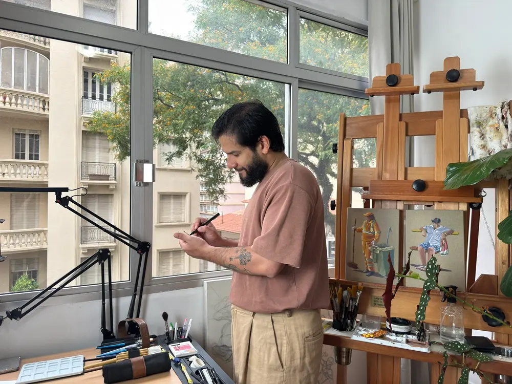 Na imagem, um artista barbudo de meia-idade, vestindo uma camiseta marrom-rosada e calças cáqui largas, está de pé em um estúdio bem iluminado, de frente para uma grande janela. Ele está olhando para baixo e segurando um instrumento de desenho ou pincel. A janela oferece uma vista para a rua, mostrando um edifício clássico de vários andares com varandas em estilo Art Déco e a folhagem verde de uma árvore. À direita, um cavalete de madeira exibe dois pequenos painéis de arte, ambos representando figuras históricas em uniformes coloridos. A mesa de trabalho em primeiro plano está cheia de materiais de arte, incluindo pincéis, paletas, e uma luminária articulada preta.