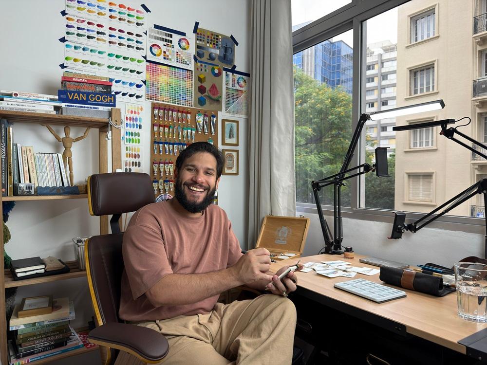Na imagem, um artista barbudo, com expressão alegre e sorriso, sentado em uma cadeira de escritório marrom, atrás de uma mesa em seu estúdio. Ele veste uma camiseta marrom-rosada e calças cáqui largas, e segura um pincel e um celular. O estúdio é bem iluminado, com uma grande janela à direita que mostra a vista de prédios urbanos e folhagem de árvores. A parede atrás do artista está decorada com gráficos de cores, cartazes de misturas de tintas e referências visuais de arte. Uma estante de madeira à esquerda está cheia de livros de arte e um pequeno manequim articulado. A mesa de trabalho contém diversos materiais de arte, como uma paleta de plástico branco, um estojo de madeira, e luminárias articuladas com luz de LED.