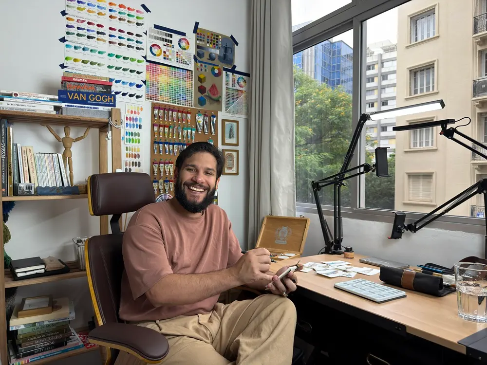 Na imagem, um artista barbudo, com expressão alegre e sorriso, sentado em uma cadeira de escritório marrom, atrás de uma mesa em seu estúdio. Ele veste uma camiseta marrom-rosada e calças cáqui largas, e segura um pincel e um celular. O estúdio é bem iluminado, com uma grande janela à direita que mostra a vista de prédios urbanos e folhagem de árvores. A parede atrás do artista está decorada com gráficos de cores, cartazes de misturas de tintas e referências visuais de arte. Uma estante de madeira à esquerda está cheia de livros de arte e um pequeno manequim articulado. A mesa de trabalho contém diversos materiais de arte, como uma paleta de plástico branco, um estojo de madeira, e luminárias articuladas com luz de LED.