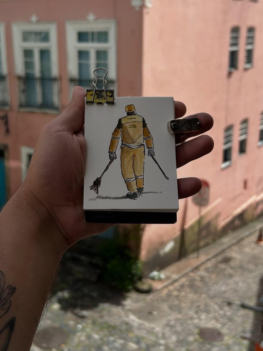 Na imagem, mão segurando um pequeno bloco de desenho ou caderno aberto, preso por dois clipes. O desenho, feito em aquarela ou guache, retrata um gari de costas, vestido com uniforme de trabalho laranja e marrom, caminhando e segurando duas vassouras nas mãos. Ao fundo, desfocado, vê-se uma rua de paralelepípedos e prédios de cores claras, possivelmente rosa antigo ou pêssego, com janelas e sacadas.