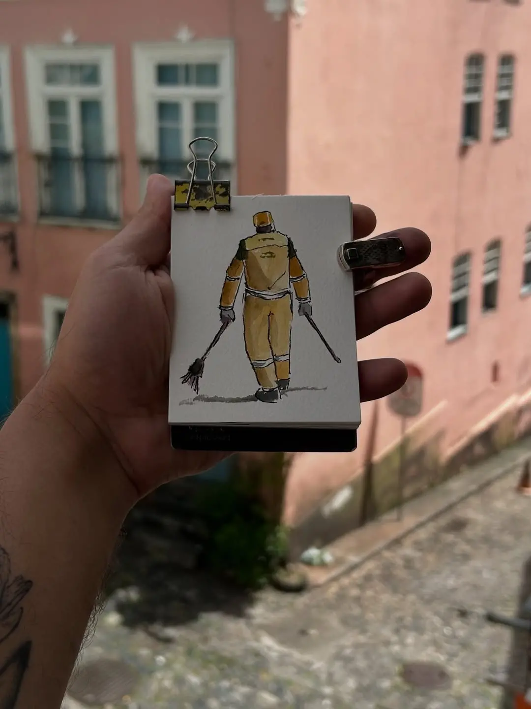 Na imagem, mão segurando um pequeno bloco de desenho ou caderno aberto, preso por dois clipes. O desenho, feito em aquarela ou guache, retrata um gari de costas, vestido com uniforme de trabalho laranja e marrom, caminhando e segurando duas vassouras nas mãos. Ao fundo, desfocado, vê-se uma rua de paralelepípedos e prédios de cores claras, possivelmente rosa antigo ou pêssego, com janelas e sacadas.