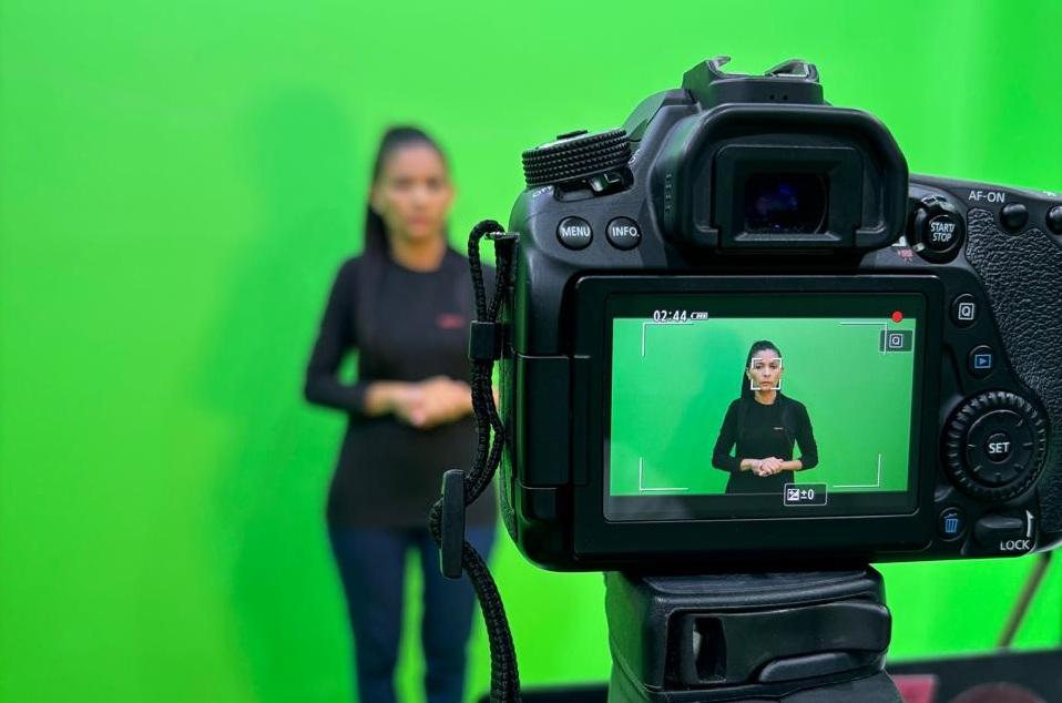 Close-up da tela de uma câmera de vídeo gravando uma mulher intérprete de Libras em frente a um fundo chroma key verde. A imagem na tela da câmera está em foco, enquanto a mulher no estúdio, no plano de fundo, está desfocada.