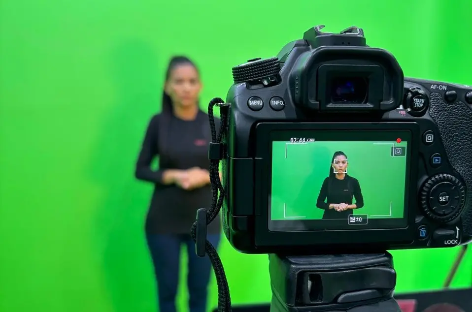 Close-up da tela de uma câmera de vídeo gravando uma mulher intérprete de Libras em frente a um fundo chroma key verde. A imagem na tela da câmera está em foco, enquanto a mulher no estúdio, no plano de fundo, está desfocada.