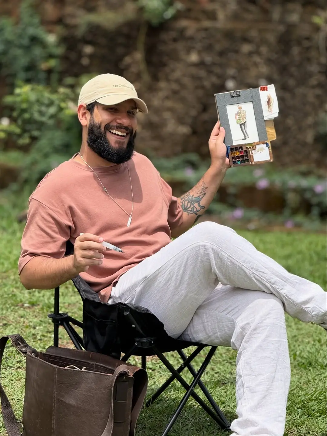 Na imagem, um homem barbudo e sorridente, com uma camiseta cor-de-rosa e calças claras, está sentado em uma cadeira de acampamento em um gramado. Ele usa um boné bege. Em sua mão esquerda, ele segura um pequeno caderno de esboços aberto, mostrando uma ilustração de figura humana. Na sua mão direita, ele segura uma caneta ou pincel. Uma bolsa de couro marrom está ao seu lado esquerdo. Ao fundo, há folhagem verde escura e um barranco gramado.