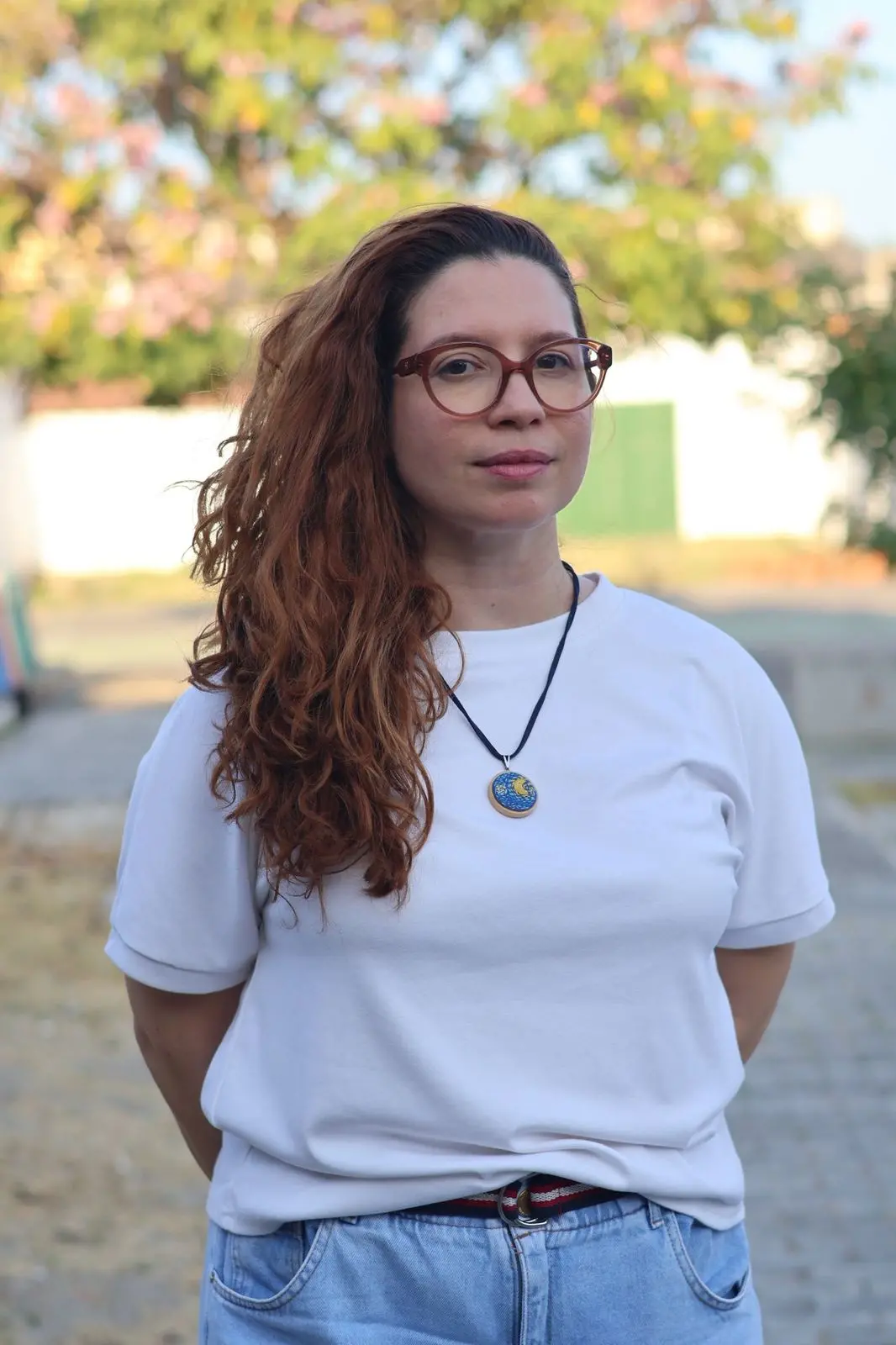 Foto de Sara Benvenuto, uma mulher branca de cabelos longos, ondulados e ruivos, usando óculos de armação marrom-avermelhada, camiseta branca básica e calça jeans. Ela está em um ambiente externo, olhando diretamente para a câmera, com uma expressão séria. Ao fundo, árvores e folhagens desfocadas em tons de verde e rosa. Ela usa um colar com um pingente redondo, azul e amarelo.