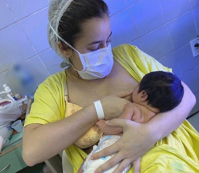 Foto em ambiente hospitalar, com luz azulada. Uma mãe jovem, usando máscara e touca descartável, segura seu bebê recém-nascido no colo, que está de fralda. A mãe veste uma camisa amarela e está amamentando o bebê. Ela tem uma pulseira de identificação hospitalar no pulso.