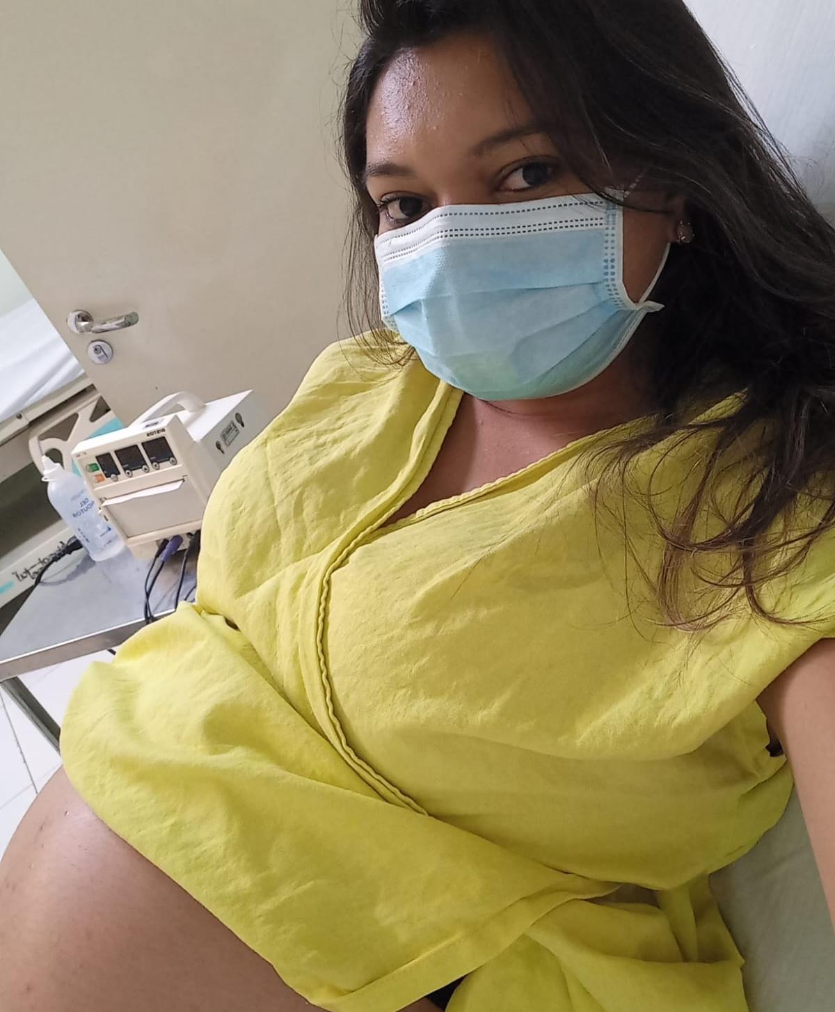 A gestante tira uma selfie em um ambiente hospitalar, provavelmente em 2021. Ela veste um roupão amarelo e está deitada, deixando a barriga em evidência. Usa uma máscara cirúrgica azul. Ao fundo, uma mesa metálica com equipamentos hospitalares e uma porta branca.