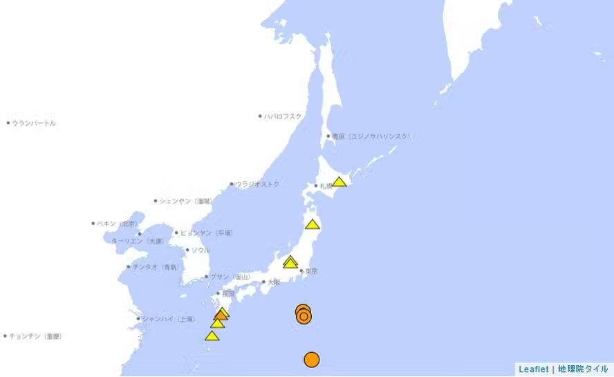 Mapa simplificado do Japão e áreas próximas do Pacífico Oeste, mostrado em fundo claro, com o contorno das ilhas japonesas destacado. Diversos ícones triangulares amarelos aparecem distribuídos sobre o território japonês — especialmente ao longo da costa leste e no centro do país — representando vulcões ativos. Mais ao sul, no oceano, há círculos concêntricos em tons de laranja, indicando a localização de terremotos recentes. Os círculos maiores sugerem eventos de maior magnitude. O mapa exibe ainda nomes de cidades e regiões em caracteres japoneses, como Tóquio, Osaka, Sapporo e outras.