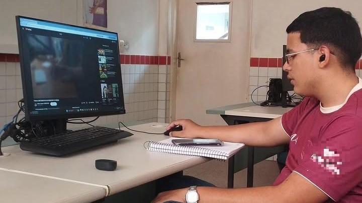 Jovem sentado em uma sala de aula usando computador de mesa. Ele veste camiseta bordô, calça jeans e chinelos, e escreve em um caderno enquanto observa a tela. A sala tem paredes claras, janelas altas e mesas com equipamentos eletrônicos.
