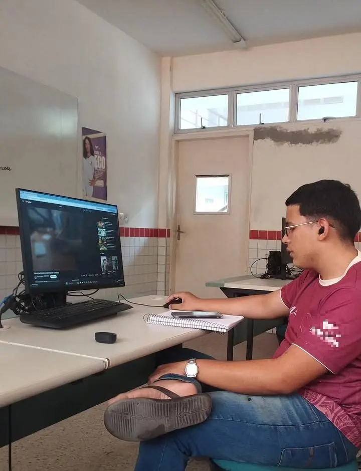 Jovem sentado em uma sala de aula usando computador de mesa. Ele veste camiseta bordô, calça jeans e chinelos, e escreve em um caderno enquanto observa a tela. A sala tem paredes claras, janelas altas e mesas com equipamentos eletrônicos.