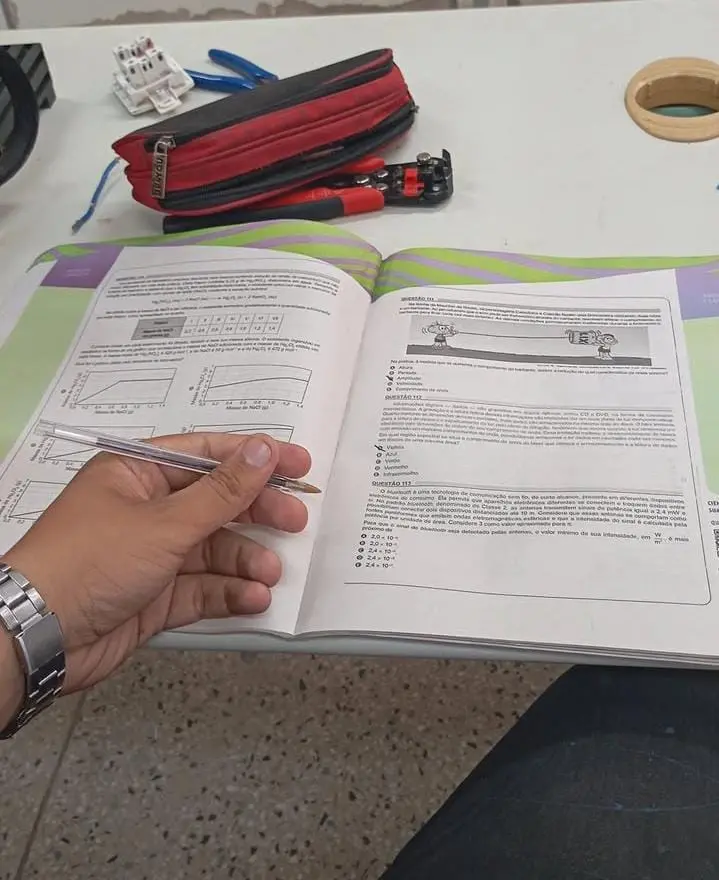 Mão de uma pessoa segurando uma caneta enquanto estuda um livro aberto com gráficos e exercícios. Sobre a mesa há um estojo vermelho, ferramentas e materiais de aula.