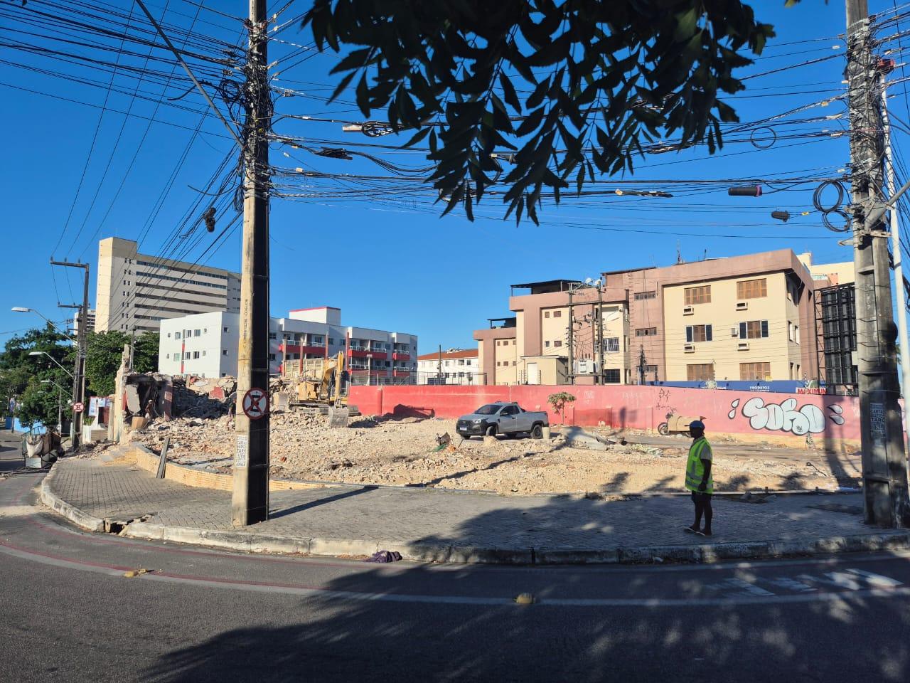 Terreno em processo de demolição cercado por entulhos, com escavadeira e caminhonete no local. Prédios residenciais aparecem ao fundo e um trabalhador com colete refletivo observa a área, sob fiação elétrica exposta e céu azul.