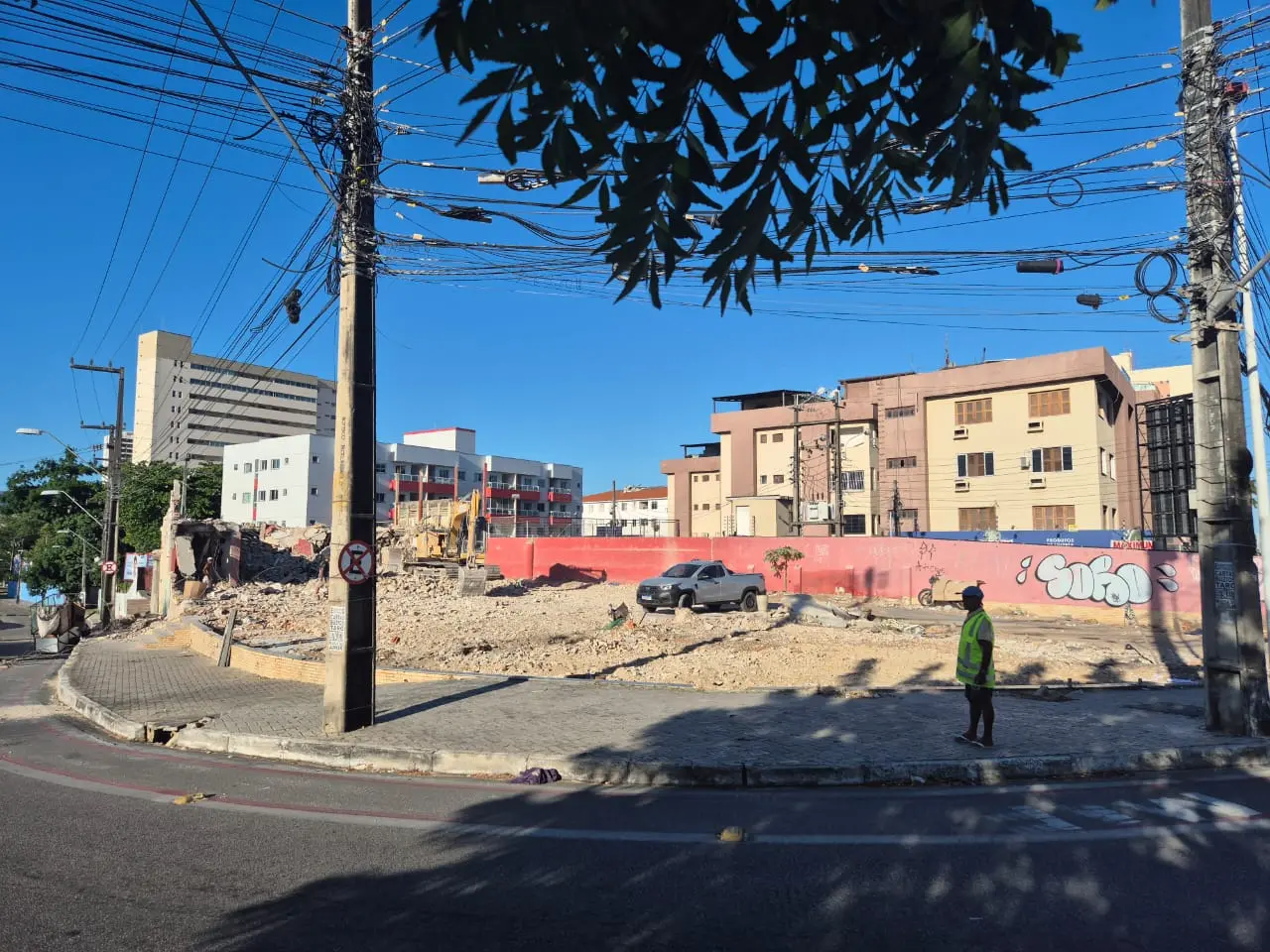 Terreno em processo de demolição cercado por entulhos, com escavadeira e caminhonete no local. Prédios residenciais aparecem ao fundo e um trabalhador com colete refletivo observa a área, sob fiação elétrica exposta e céu azul.
