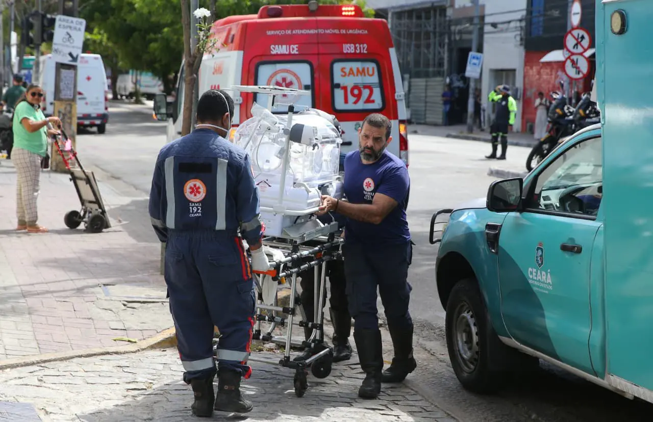 Dois profissionais do SAMU transportam uma incubadora neonatal em uma maca na calçada, ao lado de uma ambulância vermelha do SAMU 192 e de um veículo oficial do Governo do Ceará. A cena ocorre em uma rua movimentada, com pedestres e agentes de trânsito ao fundo.