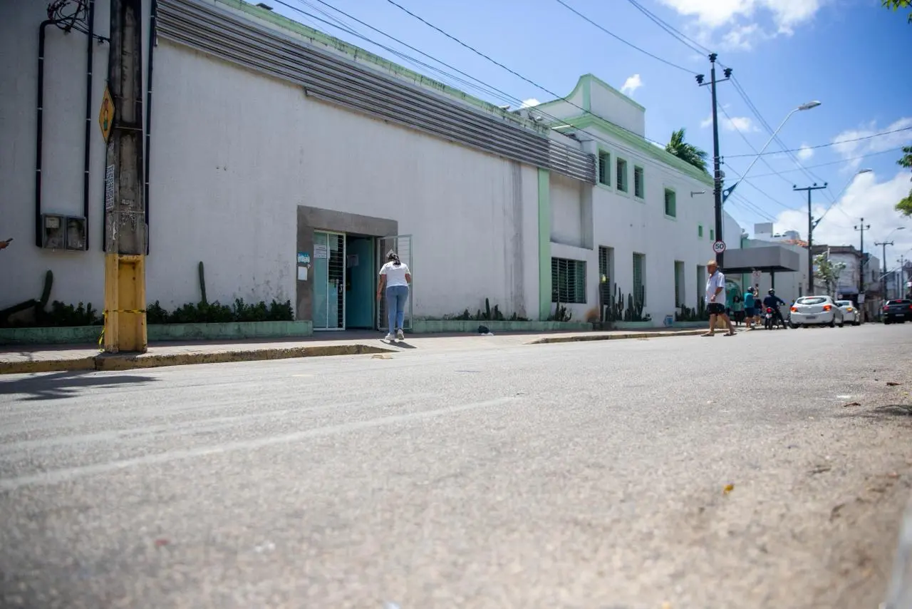 Vista externa de um prédio hospitalar de fachada clara, com janelas gradeadas e detalhes em verde. Uma pessoa caminha em direção à entrada principal, enquanto outras atravessam a rua ao fundo. O céu está azul e a rua em frente ao prédio tem pouco movimento de carros.