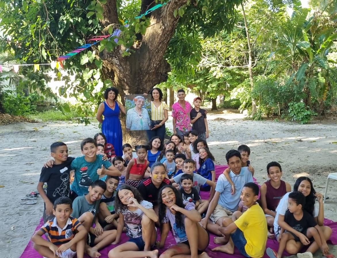 Um grupo grande de crianças e alguns adultos posam juntos, sorrindo para a câmera, sentados em uma grande lona roxa no chão sob a sombra de uma árvore, em um ambiente externo com muita vegetação. Os adultos seguram um retrato de uma senhora idosa, a artista Nice Firmeza.