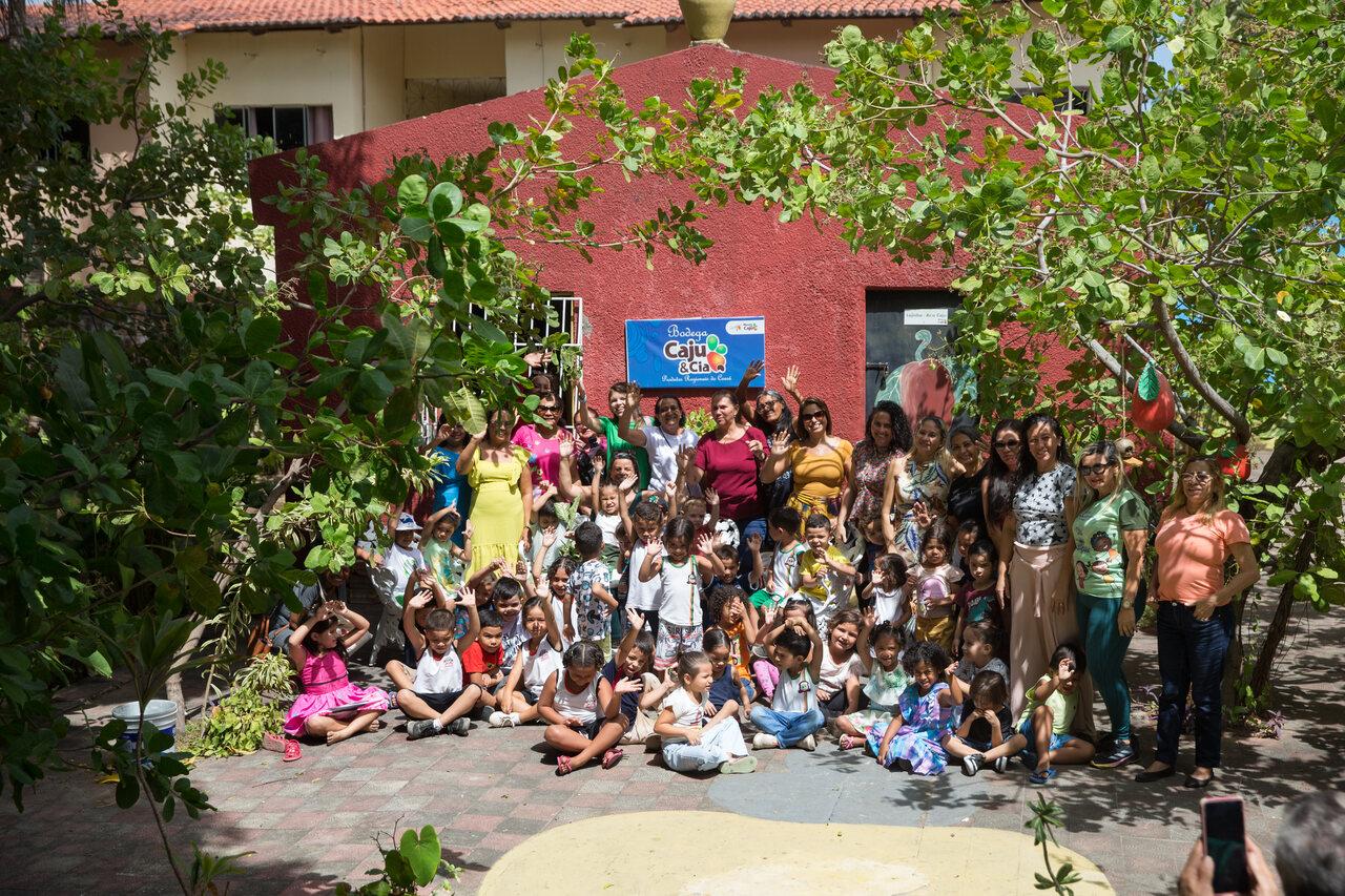 Um grande grupo de crianças e mulheres adultas posam juntas, sorrindo e acenando, em frente a um prédio vermelho, ladeado por intensa vegetação e sol forte.