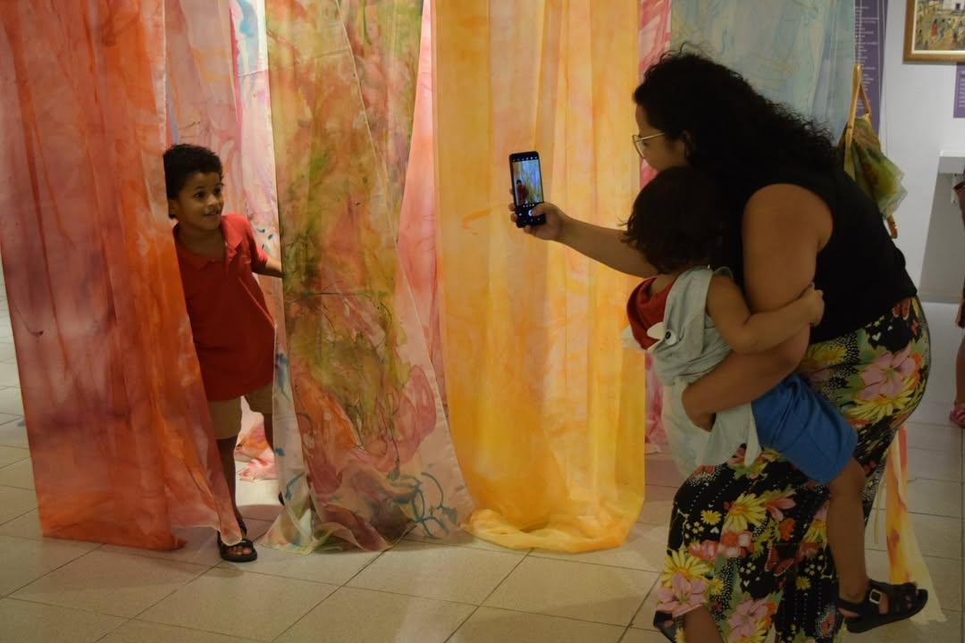 Mãe tira foto com celular de uma criança em seu colo ao lado de outra criança sorrindo, em meio a painéis de tecido transparente e colorido pendurados em uma instalação de museu.