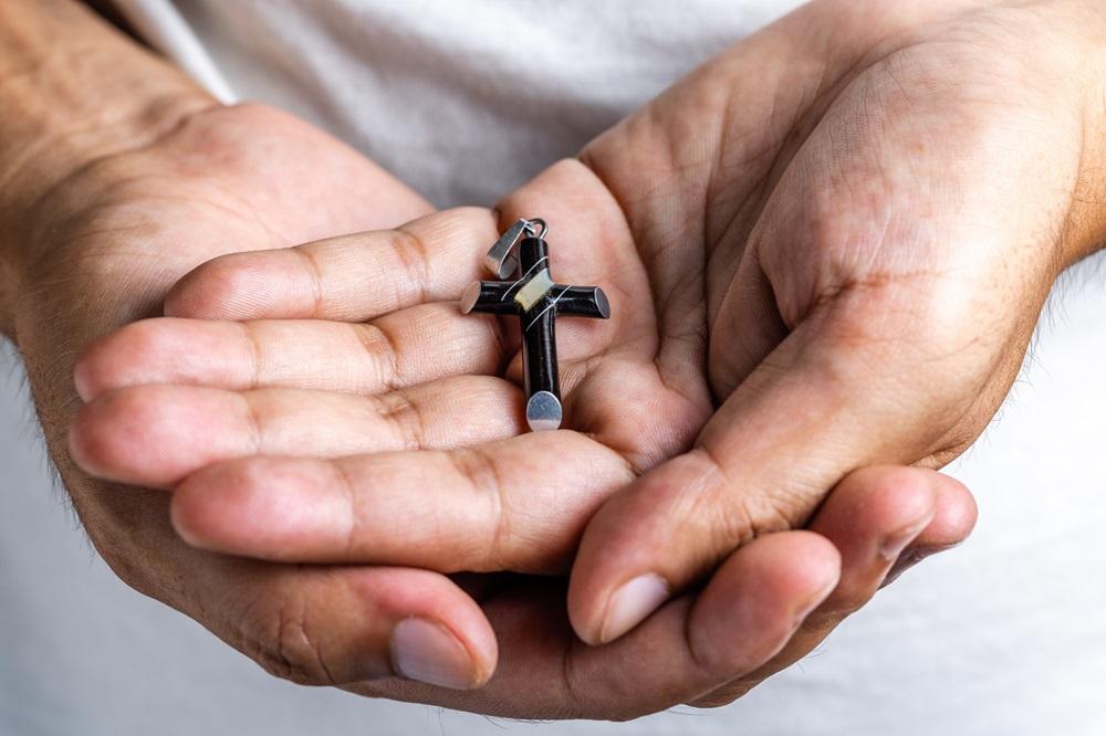 Na imagem, close-up das palmas das mãos de uma pessoa (provavelmente um homem), em formato de concha e juntas, segurando um pequeno crucifixo ou pingente de cruz preta e prateada. O crucifixo está posicionado centralmente nas palmas. O fundo está desfocado e é de um tecido branco, que parece ser a camiseta da pessoa.